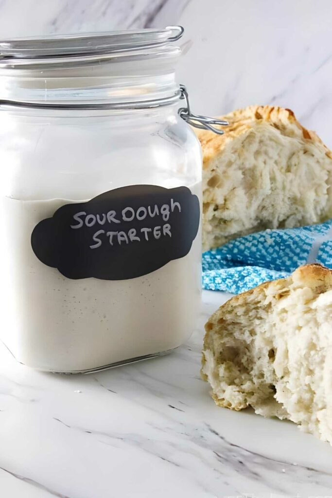 Glass jar of sourdough starter beside sliced bread and blue cloth.