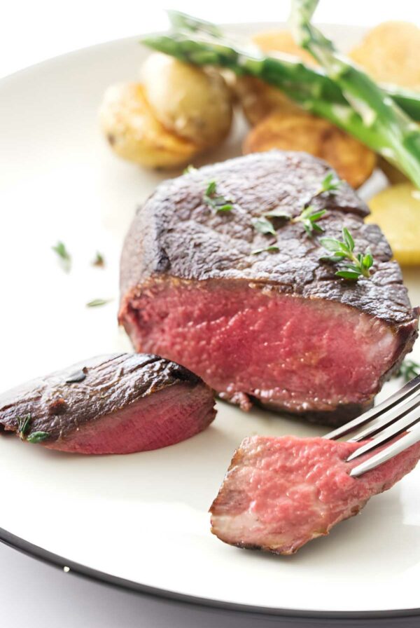 Medium-rare filet steak with herbs, potatoes, asparagus; fork holding sliced piece.