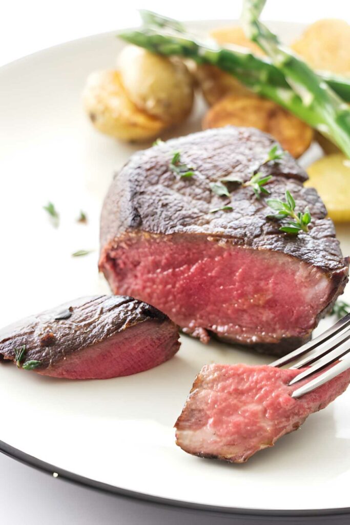 Medium-rare steak with herbs, potatoes, asparagus; fork holding sliced piece.