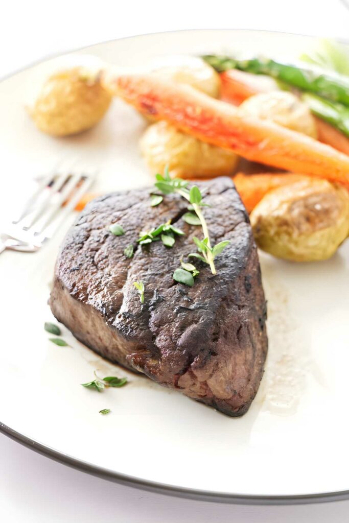 Cooked steak with herbs, roasted potatoes, carrots, and asparagus on plate.