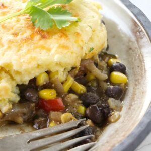 Close-up of cornbread-topped casserole with beans, corn, peppers, parsley, and fork.