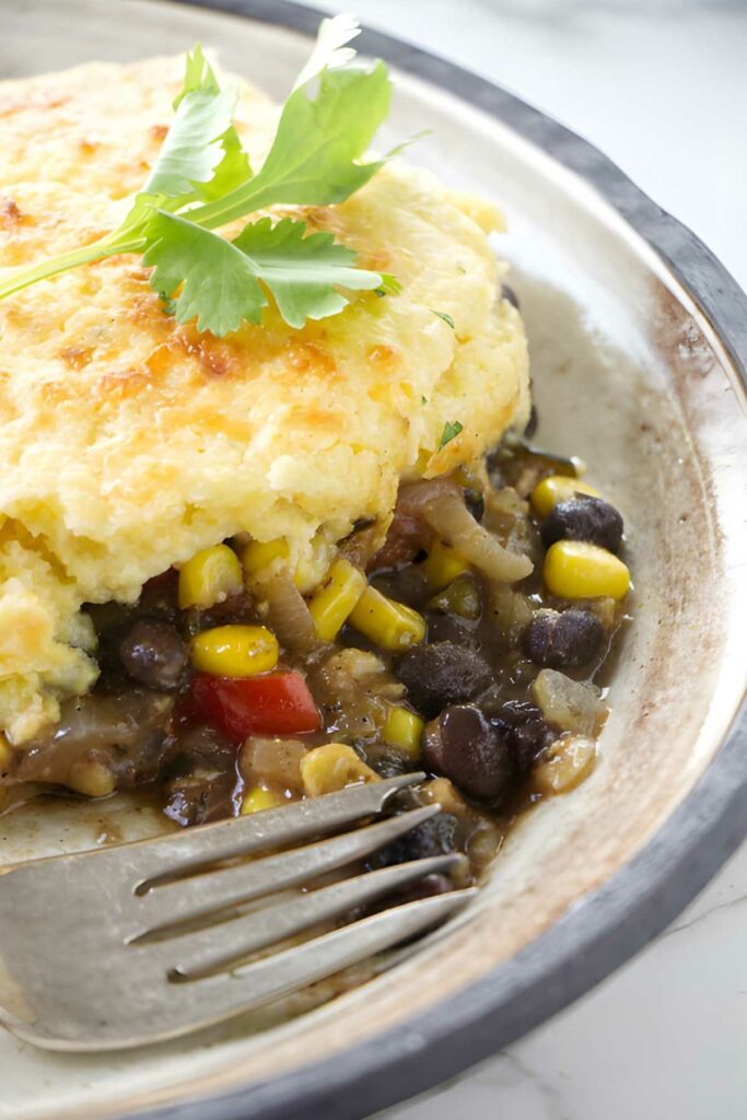 Close-up of cornbread-topped casserole with beans, corn, peppers, parsley, and fork.