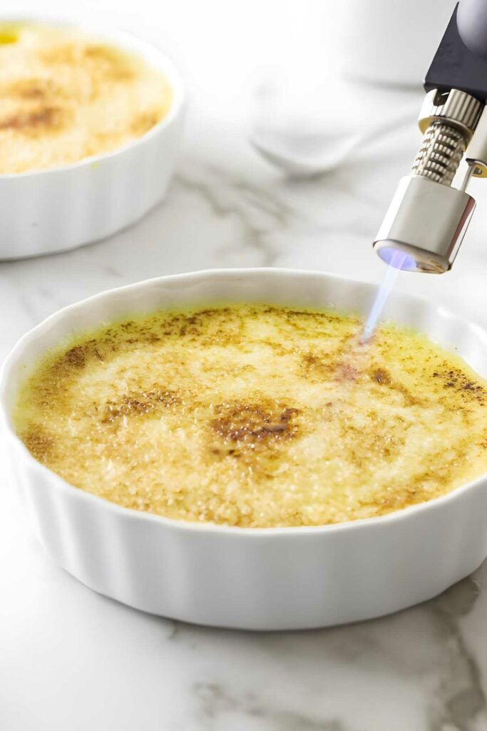 Kitchen torch caramelizes crème brûlée in white ramekin on marble surface.