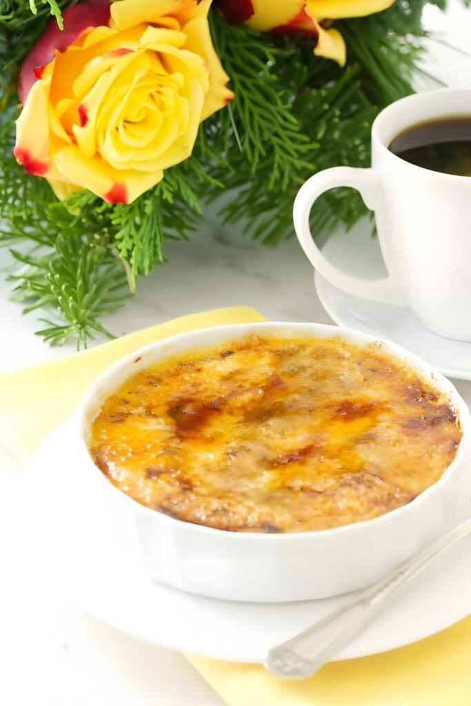 Crème brûlée in a white ramekin with spoon, coffee, and yellow roses nearby.
