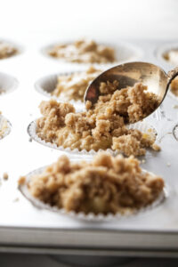 A spoon adding streusel topping to muffins in a muffin tin.