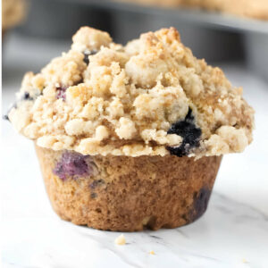 A blueberry muffin with a crumble topping in front of a muffin tin.