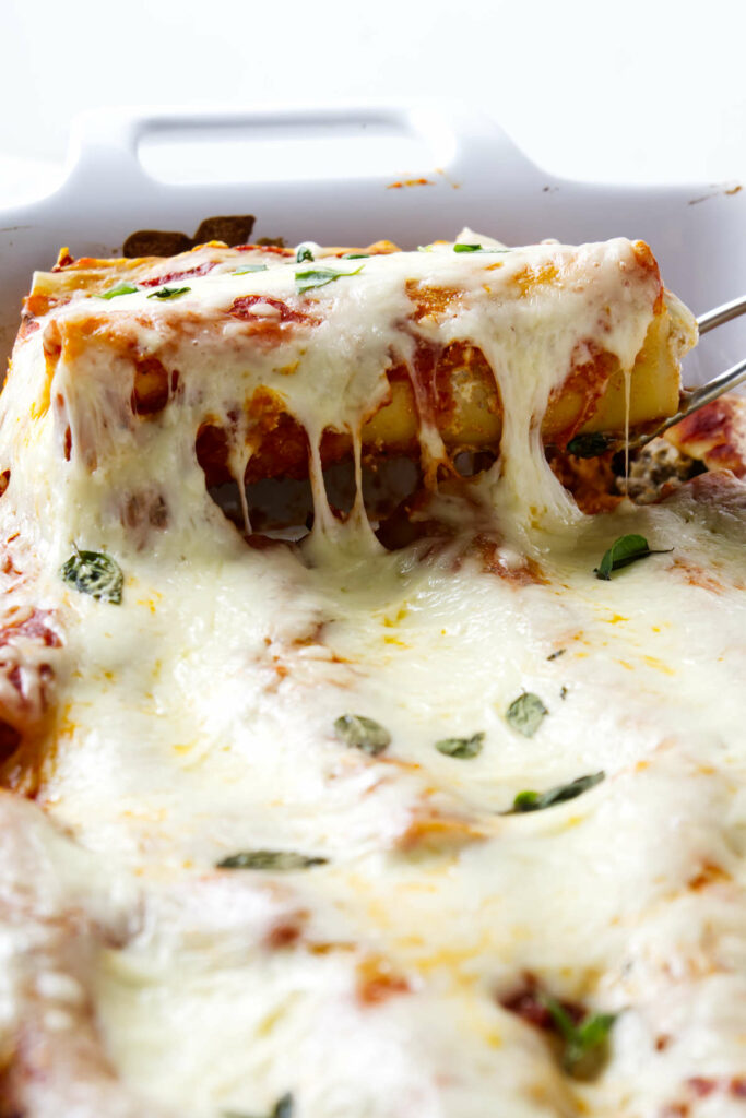 A spatula lifting a serving of hot cannelloni pasta tubes from a casserole dish.