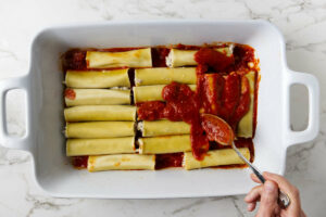Spreading marinara sauce on top of filled canneloni pasta.
