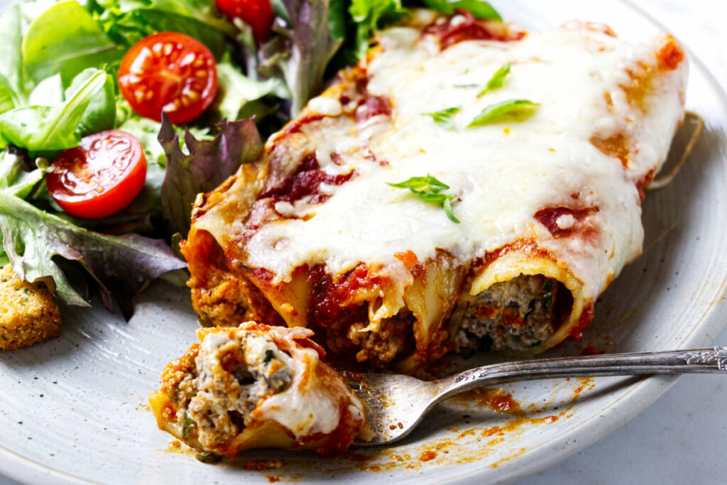 A fork next to three beef filled cannelloni on a plate with a salad.