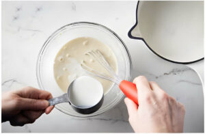 Pouring hot milk into the egg mixture to temper the egg yolks.