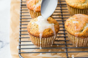 Drizzling lemon glaze over the top of the warm muffins on a cooling rack.