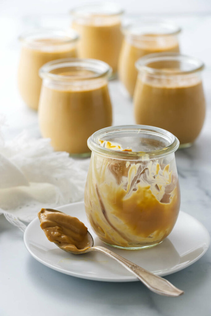 Six jars of custard caramel pudding next to a spoon.