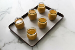 Six jars of caramel custard on a baking sheet.