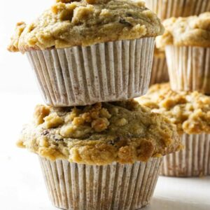 Two crumb-topped muffins stacked, more muffins blurred in the background.