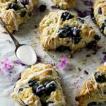 Glazed blueberry scones on parchment with spoon and pink flowers nearby.