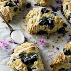 Glazed blueberry scones on parchment with spoon and pink flowers nearby.