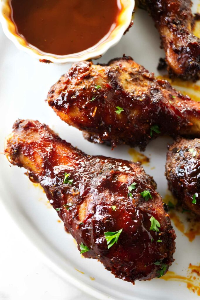 Barbecued chicken drumsticks with parsley on a plate, barbecue sauce behind.