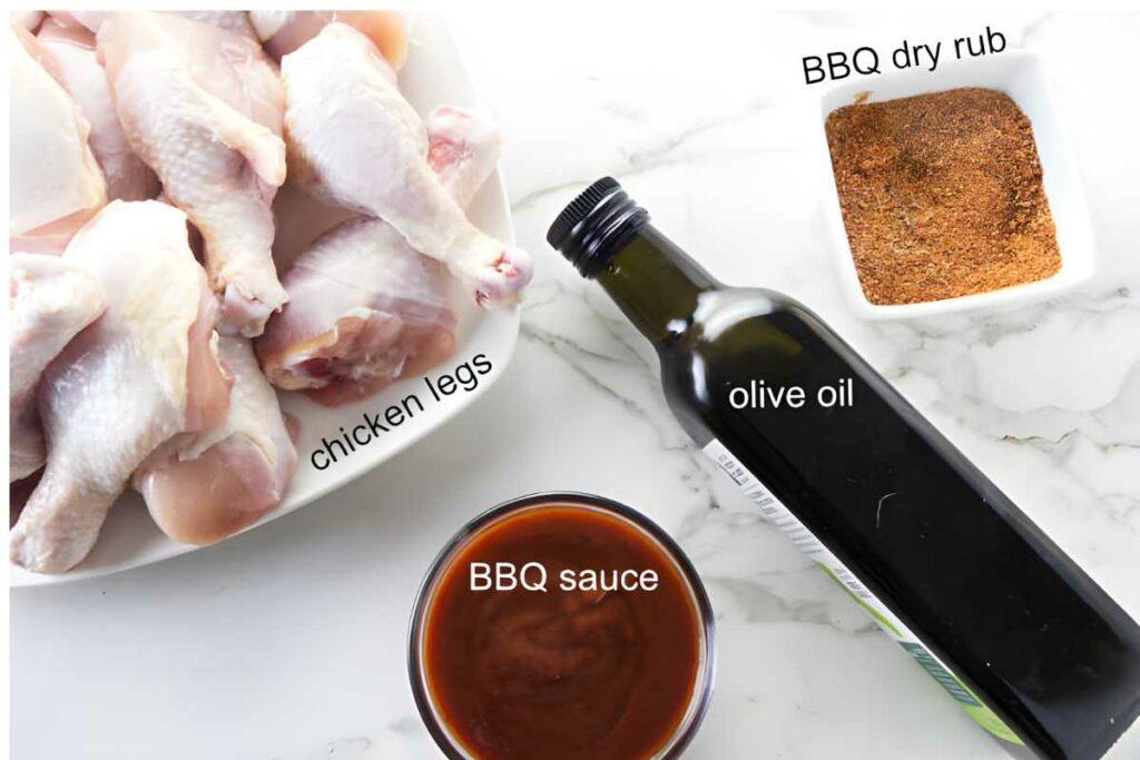 Raw chicken legs, BBQ sauce, olive oil, and dry rub on labeled marble surface.