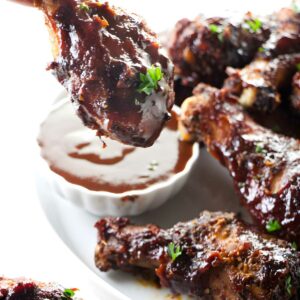 Hand holding glazed chicken drumstick over barbecue sauce, with saucy chicken pieces.