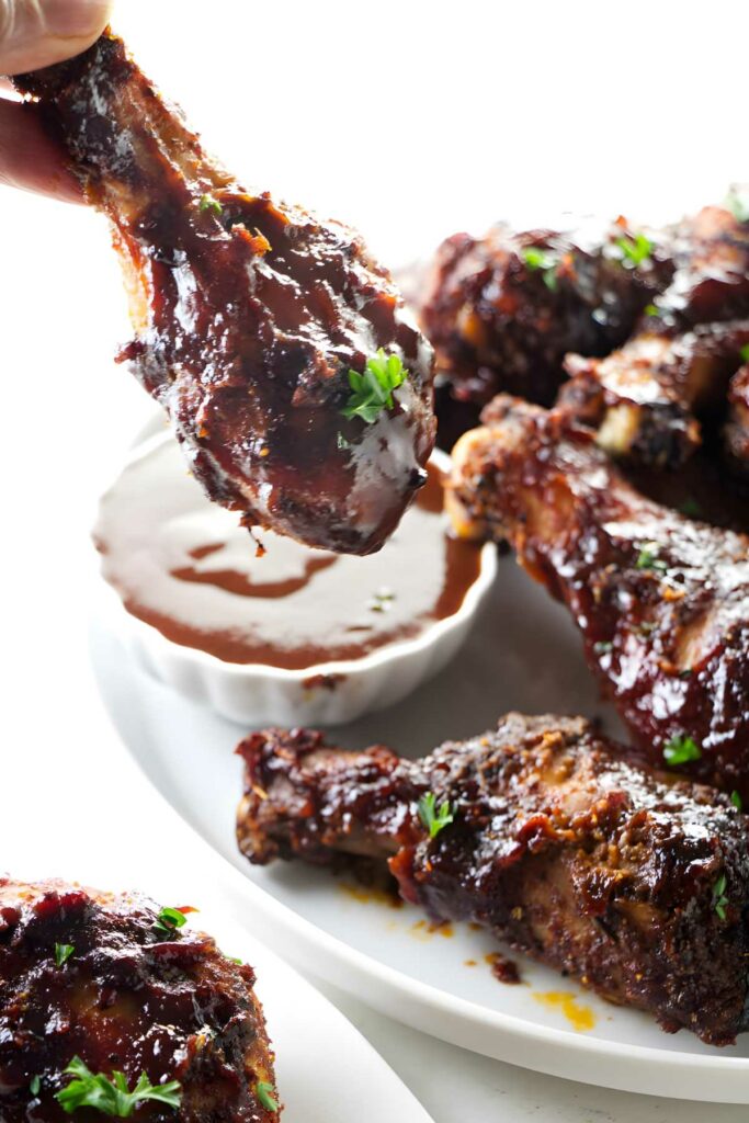 Hand holding glazed chicken drumstick over barbecue sauce, with saucy chicken pieces.