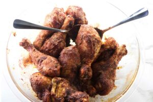 Seasoned chicken drumsticks in a glass bowl with metal serving spoons.