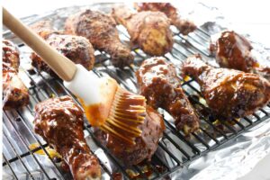 Hand brushing barbecue sauce on grilled chicken drumsticks on foil-lined wire rack.