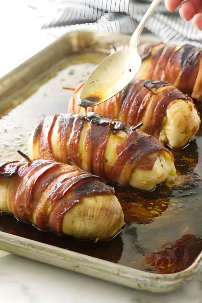 Bacon wrapped chicken breasts on a baking sheet, sauce being spooned over one.