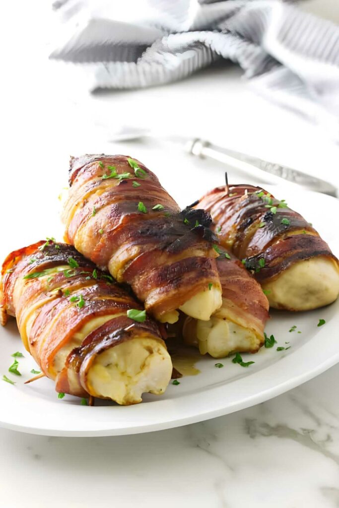 Bacon wrapped, cheese-stuffed chicken breasts on a white plate with herbs.