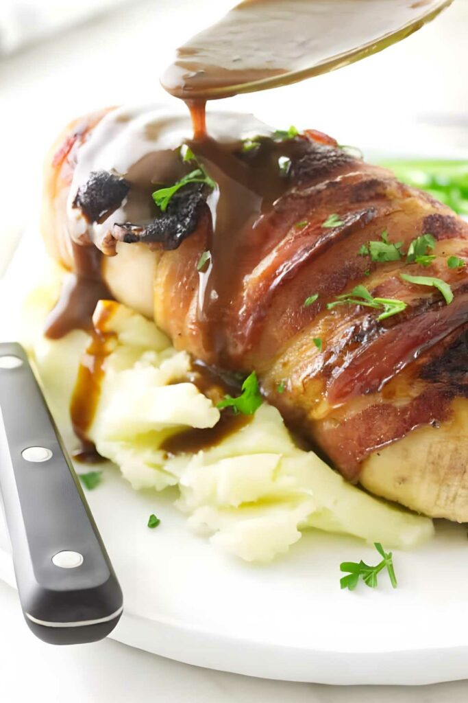 Bacon-wrapped chicken on mashed potatoes with gravy and parsley garnish.