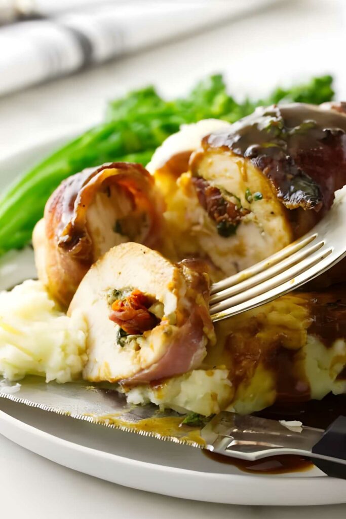 Fork slices bacon-wrapped stuffed chicken on mashed potatoes with gravy and greens.