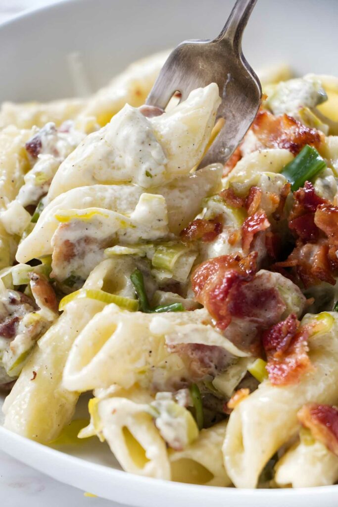 Creamy penne pasta with bacon, leeks, and white sauce on a fork.