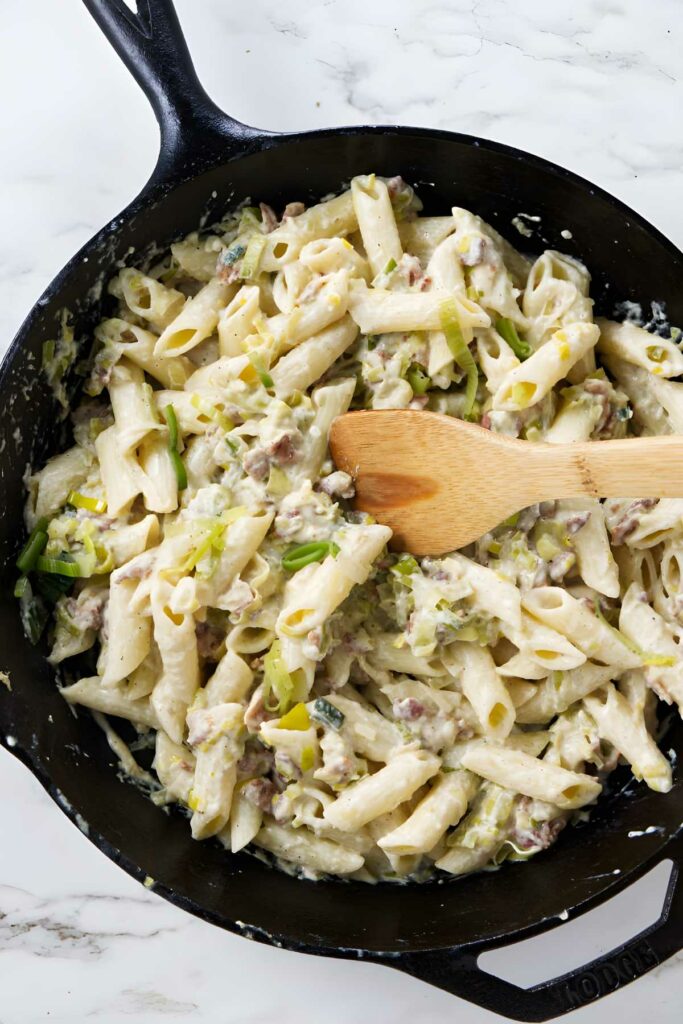 A skillet with a spoon stirring pasta with bacon and leeks. 