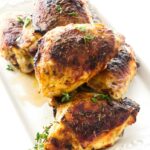 Roasted chicken thighs with crispy skin on a white platter, garnished with thyme.
