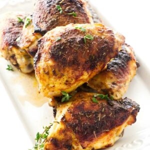 Roasted chicken thighs with crispy skin on a white platter, garnished with thyme.