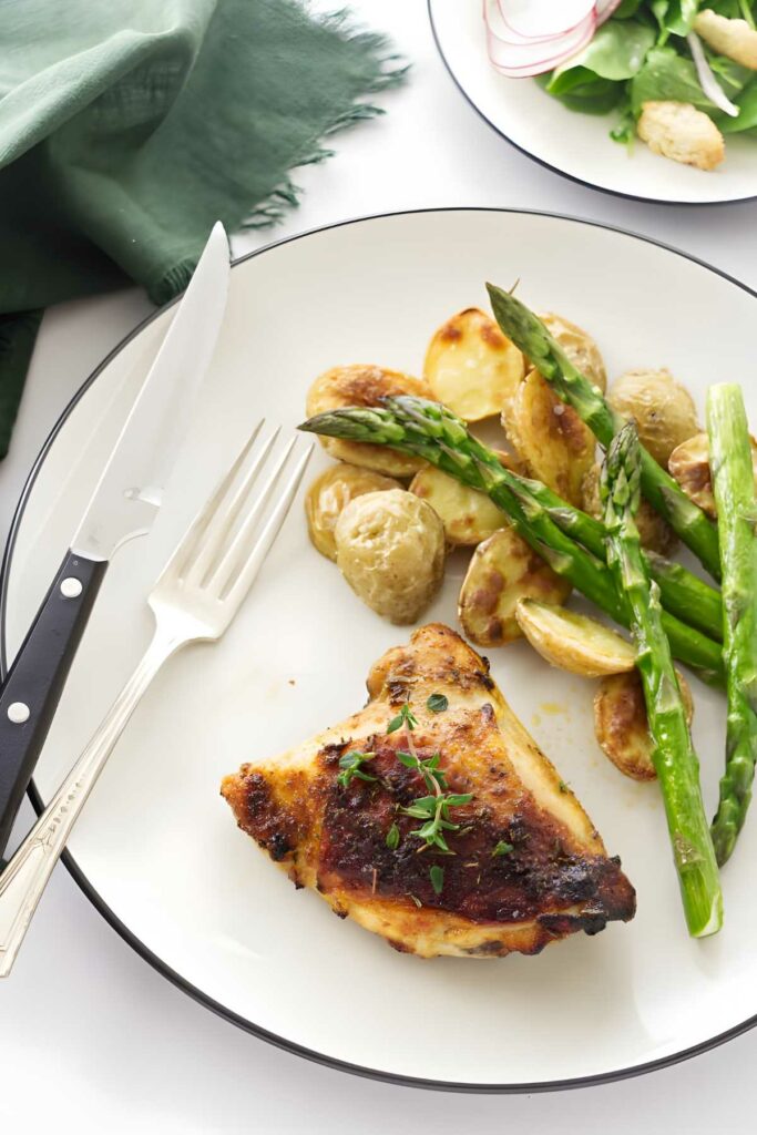 Roasted chicken thigh with potatoes and asparagus on a plate, utensils beside.