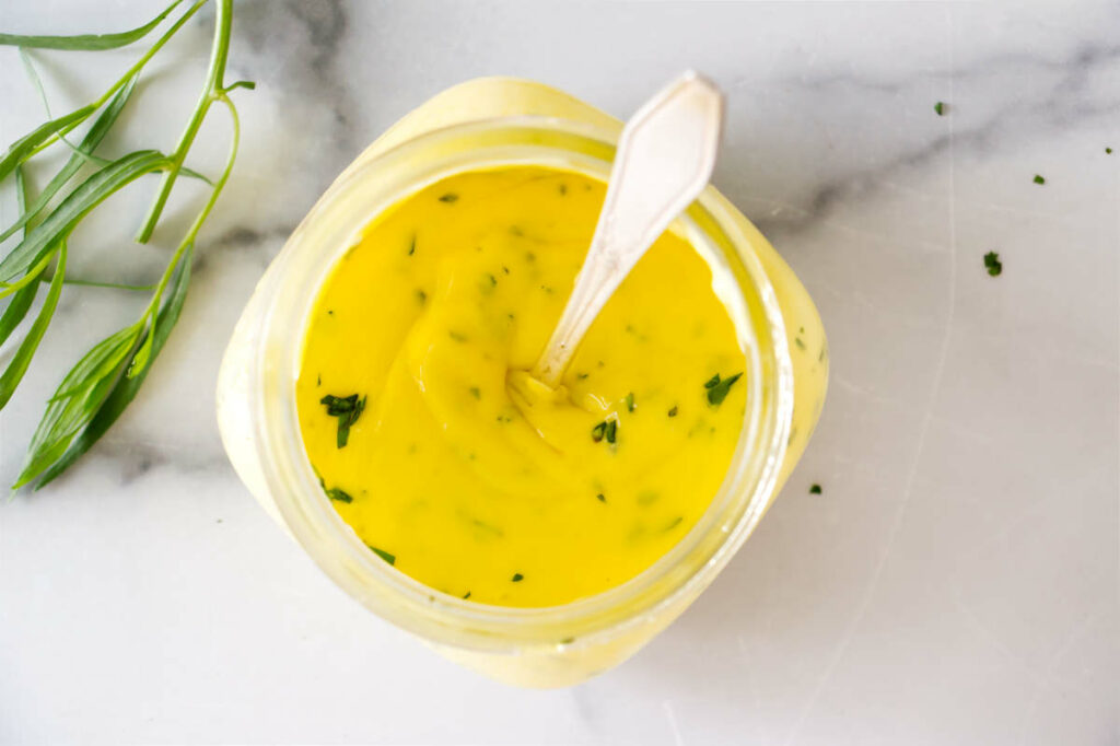 A spoon in a jar of bernaise sauce next to a sprig of tarragon.