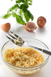 Glass bowl with beaten eggs and cheese mixed by fork, herbs behind.