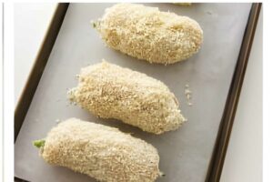 Four breaded stuffed jalapeños sit on a baking sheet, ready to bake.