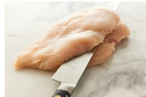 Slicing a raw chicken breast in half with a large kitchen knife.