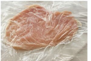 Thinly pounded raw chicken breast partially covered in wrinkled plastic wrap.