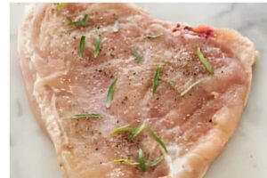 Heart-shaped raw chicken breast with herbs, salt, and pepper on light surface.