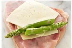 Raw chicken breast with ham, Swiss cheese, and asparagus on marble.