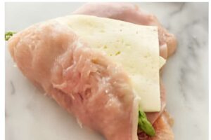 Raw chicken breast folded over white cheese and asparagus on marble.