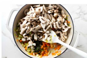White pot with chopped mushrooms, celery, carrots, and herbs being stirred.