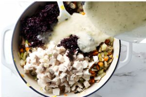 Creamy sauce is poured over chicken, vegetables, and wild rice in a pot.