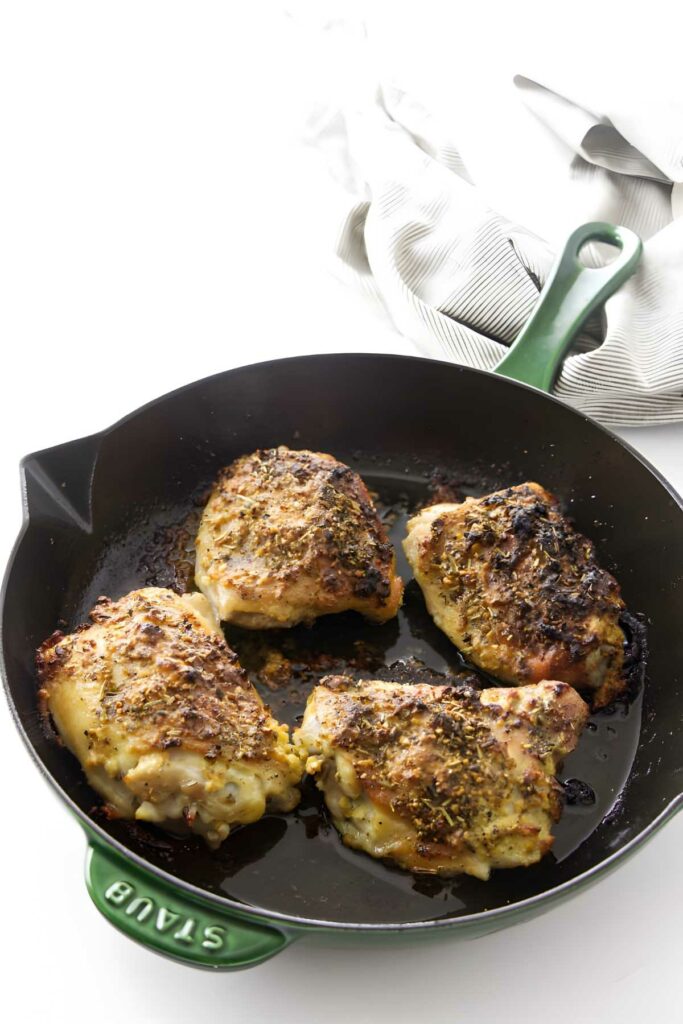 Cooked chicken thighs in black skillet with green handle on white surface.