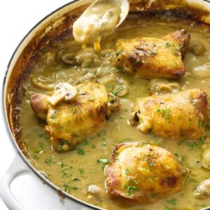 Golden-brown chicken thighs in creamy sauce in casserole, spoon pouring sauce.