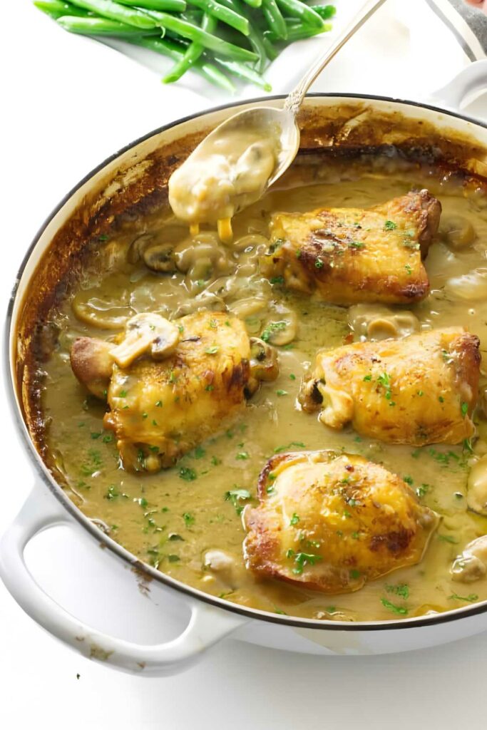 Golden-brown chicken thighs in creamy sauce in casserole, spoon pouring sauce.