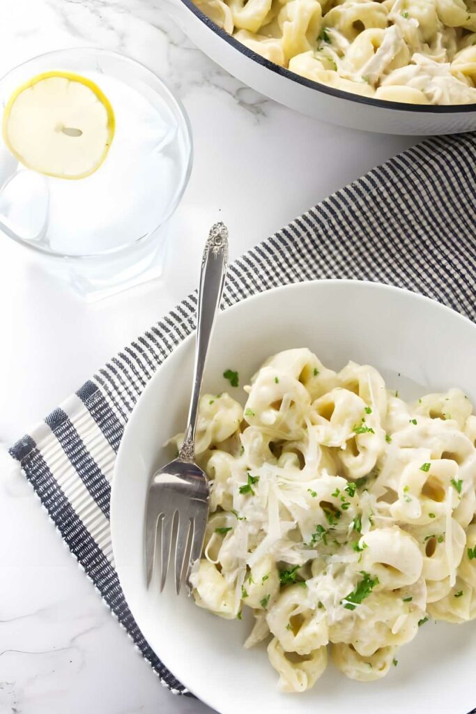 Bowl of creamy tortellini with parsley and cheese, fork and lemon water nearby.