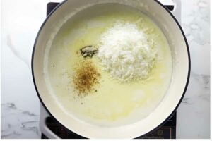 White pot with melted butter, cheese, pepper, and nutmeg on stovetop.